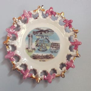 May include: A decorative ceramic plate with a white center featuring a colorful illustration of Sault Ste. Marie, Michigan. The plate has a scalloped edge with pink and gold accents. The text "SAULT STE. MARIE MICHIGAN" is visible.