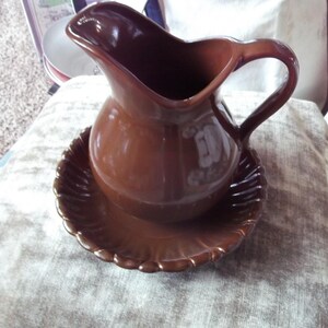May include: A brown ceramic pitcher with a handle, sitting on a matching plate. The pitcher has a rounded body and a spout for pouring. The plate has a scalloped edge. The set is likely for serving water or other beverages.