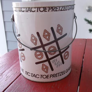 May include: White metal tin with a black and brown noughts and crosses game printed on it. The tin has a wire handle and says "Tic Tac Toe Pretzel Game" on the side.