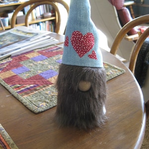 May include: A denim gnome with a brown fuzzy beard and a hat with three red hearts. The hat is made of denim and has white polka dots on the hearts.