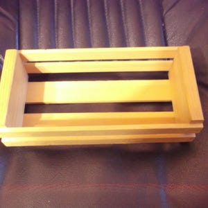 May include: A small, rectangular wooden crate with a natural finish. The crate has a slatted design with three horizontal slats and solid end pieces. The crate is empty and sits on a dark brown surface.