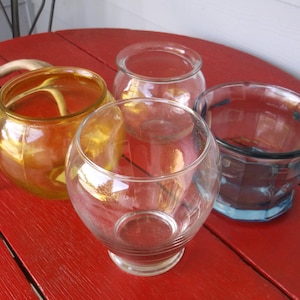 May include: Four glass bowls of varying shapes and sizes sit on a red surface. One is amber-colored, another is clear with a round shape, and the others are clear with a cylindrical shape.