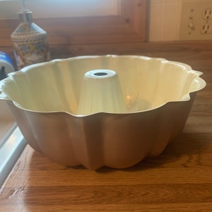 May include: A light beige bundt cake tin with a central tube and scalloped edges. The metal tin sits on a wooden countertop. A decorative soap dispenser is visible in the background.