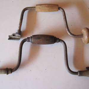 May include: Two vintage hand-crank drills with wooden handles. The drills are made of metal and have a classic design.