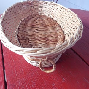 May include: A woven wicker basket with a round base and a flared, open top. The basket has a natural, light brown color with a woven handle. The basket is sitting on a red surface.