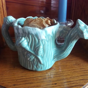 May include: A pale turquoise ceramic watering can-shaped vase with a handle and spout. The vase contains dried flowers. The vase is on a wooden surface.