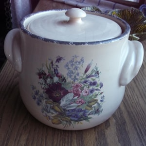 Peut inclure: Pot à biscuits en céramique crème avec couvercle et poignées latérales. Le pot est orné d'un bouquet floral aux tons violets, roses et bleus. Le couvercle a un petit bouton. Le bord est bordé d'une bordure bleue.