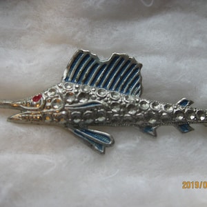 May include: A silver-toned brooch in the shape of a marlin fish. The fish is decorated with rhinestones and has blue enamel accents. The marlin's eye is a red cabochon.