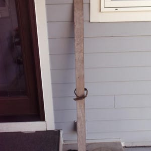 May include: A single, vintage wooden ski, standing upright against a light gray wall. The ski is a light brown color, with a pointed tip and a metal binding. The ski is weathered, suggesting age and use. A dark brown door and a window are visible in the background.