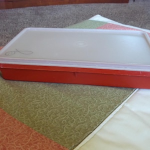 May include: A rectangular red plastic container with a clear lid. The lid has a small design on it. The container is sitting on a patterned quilt with green, red, and white sections.