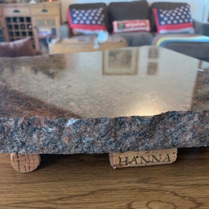 May include: A rectangular granite serving board with a polished top and rough, natural edges. The board is supported by four cork feet. The granite has a mix of brown, black, and pink tones. One cork foot has the name "HANNA" printed on it.