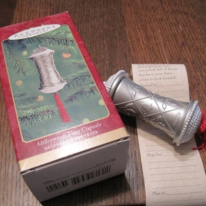 Peut inclure: Une décoration de Noël en forme de capsule temporelle du millénaire en argent avec un pompon rouge. La décoration est dans sa boîte d'origine, qui a un fond rouge et les mots "Millennium Time Capsule" et "Decoration Des Fetes" imprimés dessus.