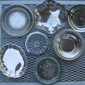 May include: A collection of seven silver-plated serving dishes of various sizes and shapes, arranged on a grey metal grid surface.