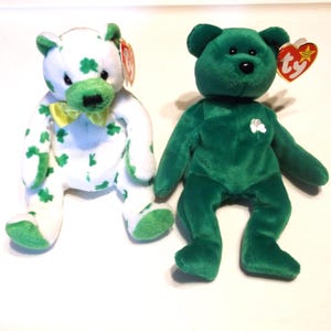 May include: Two plush teddy bears. One is white with green shamrocks and accents, a yellow bow tie, and a red tag. The other is solid green with a black nose and a red tag. Both are sitting.