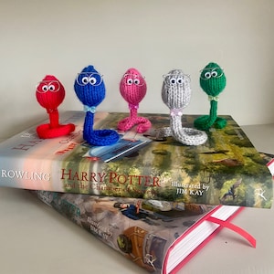 May include: Five knitted snake figurines wearing glasses, sitting on a copy of "Harry Potter and the Chamber of Secrets" by J.K. Rowling. The snakes are red, blue, pink, gray, and green.