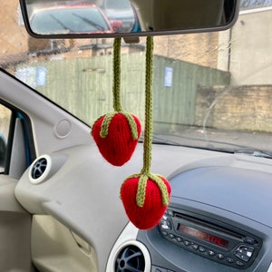 May include: Two crocheted strawberries with bright red bodies and green stems, each attached to a long green crocheted cord. The strawberries are hanging from a car rearview mirror, featuring a textured yarn construction with visible stitches.