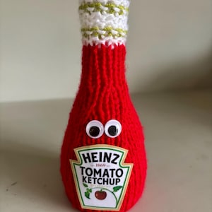 Peut inclure: Une bouteille de ketchup en tricot rouge avec des yeux globuleux et une étiquette Heinz Tomato Ketchup. La bouteille a un dessus en tricot blanc avec des rayures vertes.