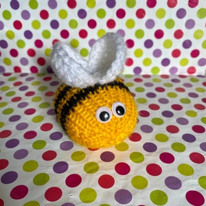 May include: A crocheted bee toy, featuring a yellow body with black stripes and white wings. The bee has googly eyes. The background is a colorful polka dot pattern. This handmade item could be a decorative piece or a child's toy.