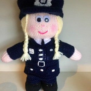 Könnte beinhalten: Eine handgestrickte Puppe in einer dunkelblauen Polizeiuniform mit passender Mütze und weißem Sternemblem. Die Puppe hat blondes Garnhaar in zwei Zöpfen, Wackelaugen und einen rosa gestickten Mund. Ein weißer Aufnäher auf der Brust trägt PS HAY.