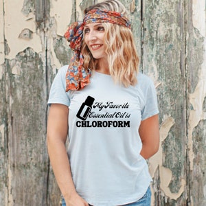 以下が含まれることがあります： 黒いボトルと「My Favorite Essential Oil is CHLOROFORM」というテキストが書かれた薄い青色のTシャツ。