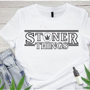 May include: White t-shirt with the text "STONER THINGS" in a black, retro-style font, featuring a cannabis leaf graphic. The shirt is paired with blue jeans and cannabis leaves.