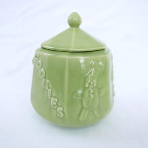 May include: A light green ceramic cookie jar with a lid. The jar has the word "COOKIES" in raised letters on the side, along with a raised image of a gingerbread man chef. The jar is a light green color.