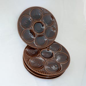 May include: A stack of brown ceramic oyster plates. Each plate has seven circular indentations and a central well. The plates have a ridged, textured rim and a glossy finish. The plates are stacked on a white surface.