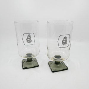 May include: Two clear glass drinking glasses with a square base. Each glass has a gray logo with the word "ZUIDER" and a ship design.
