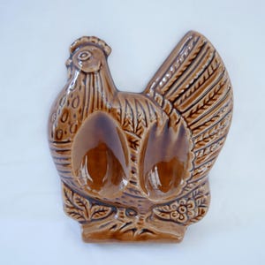 May include: Brown ceramic rooster-shaped spoon rest. The spoon rest features a detailed design with a textured body, a comb, and a tail. The spoon rest has two spoon holders and is decorated with floral accents.