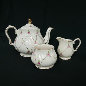 May include: A white porcelain tea set comprising a teapot, sugar bowl, and creamer. The set is decorated with pink roses, gold trim, and accents. The teapot has a gold handle and lid finial.