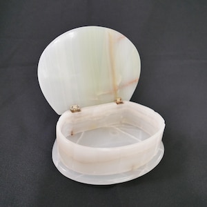 May include: An open, oval-shaped onyx box with a hinged lid. The box is white with green and tan veining. The lid is open, revealing the interior of the box. The box sits on a black surface.