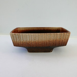 Pode incluir: Uma tigela retangular de cerâmica com interior e exterior marrom. O exterior tem um design texturizado, com nervuras verticais com um gradiente de tons marrons e bege. A tigela fica em uma pequena base marrom escuro.