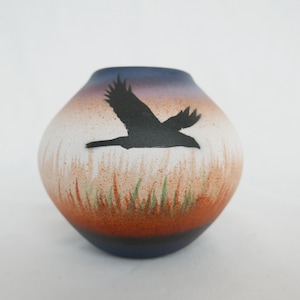 Peut inclure: Petit vase rond en céramique avec un motif peint. Le vase présente un oiseau noir en vol sur un fond de coucher de soleil orange, blanc et bleu. De l'herbe verte est peinte au bas du vase.