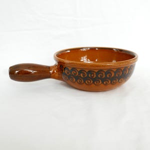 Op de afbeelding: Een bruine keramische kom met een handvat. De kom heeft een donkerbruine rand en een decoratief zwart wervelpatroon aan de buitenkant. Het handvat is ook bruin en lijkt van hetzelfde materiaal te zijn als de kom. De kom wordt waarschijnlijk gebruikt om voedsel te serveren.