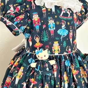 May include: A dark blue dress with a white lace collar and short sleeves. The dress features a colourful print of nutcrackers, ballerinas, and Christmas trees. A small stuffed cat with a blue bow is attached to the front.