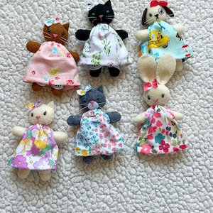 May include: A collection of handmade fabric animal dolls, including cats, a dog, and a bunny. Each doll is dressed in a colorful floral or patterned dress and has a bow. The dolls are arranged on a textured, off-white surface.