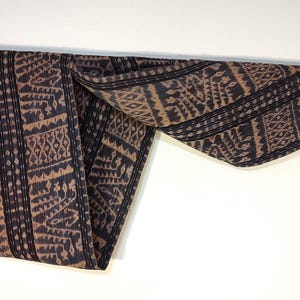 May include: A rectangular textile featuring a dark brown and black ikat pattern with repeating geometric motifs and vertical stripes. The fabric is draped over a horizontal wooden rod, displaying a complex woven design with visible frayed edges.