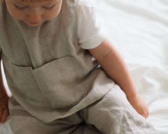 May include: A young child wearing a light brown linen jumpsuit with a pocket on the front. The child is sitting on a white surface.