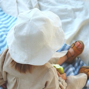 May include: A young child wearing a white linen sun hat and a beige linen dress sits on a blue and white striped blanket. The child is holding a green apple in their hand.