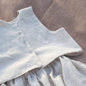 Könnte beinhalten: Ein weißes ärmelloses Oberteil mit einem gerafften Rock. Das Oberteil hat einen runden Ausschnitt und einen Knopfverschluss. Der Rock ist in der Taille gerafft und hat eine fließende Silhouette.