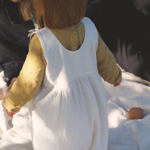 May include: A white linen pinafore dress with a scoop neck and adjustable straps. The dress has a gathered skirt and is being worn by a child.