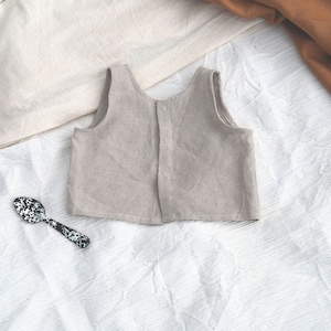 Puede incluir: Una blusa sin mangas de lino beige claro con cierre de botón en la espalda. La blusa está colocada sobre una superficie blanca texturizada.