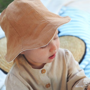 May include: A young child wearing a brown linen sun hat with a white trim and a beige linen button-up shirt.