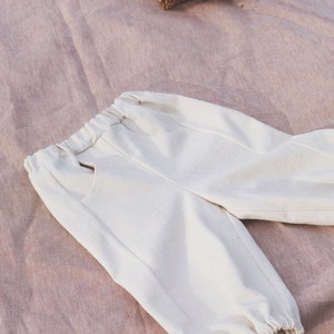 Op de afbeelding: Witte katoenen broek met een elastische tailleband en getailleerde pijpen. De broek ligt plat op een roze stoffen oppervlak.