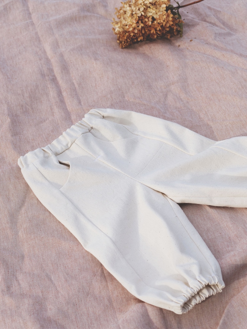 Op de afbeelding: Witte katoenen broek met een elastische tailleband en getailleerde pijpen. De broek ligt plat op een roze stoffen oppervlak.