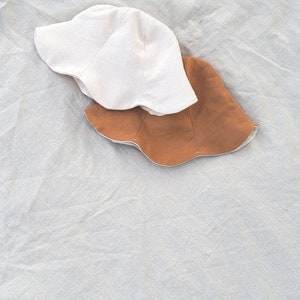 May include: Two sun hats, one white and one brown, are stacked on top of each other on a white fabric background.