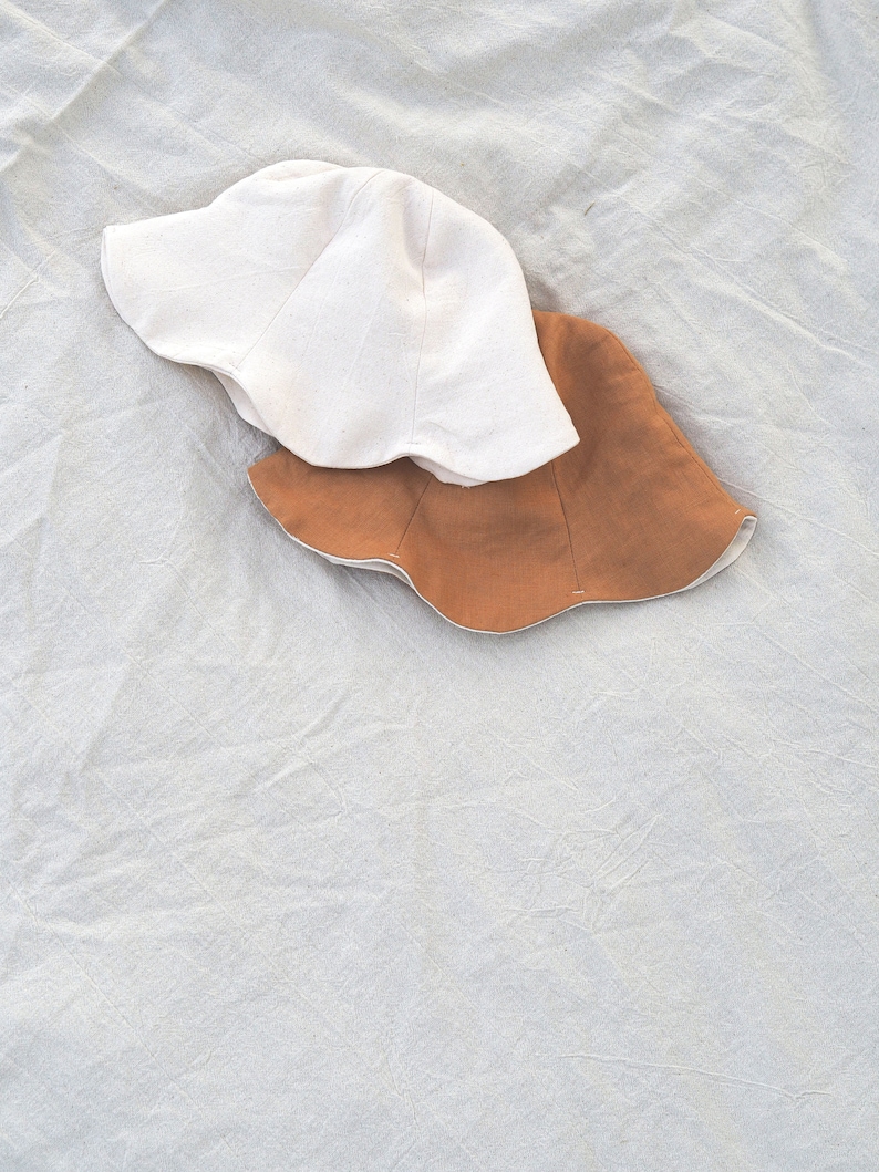 May include: Two sun hats, one white and one brown, are stacked on top of each other on a white fabric background.