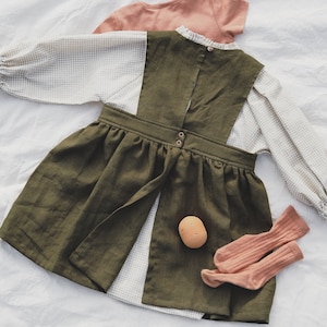 May include: A green linen dress with a white and beige gingham overlay. The dress has a gathered skirt and a button closure at the back. The dress is paired with a pair of pink socks and a small brown toy.