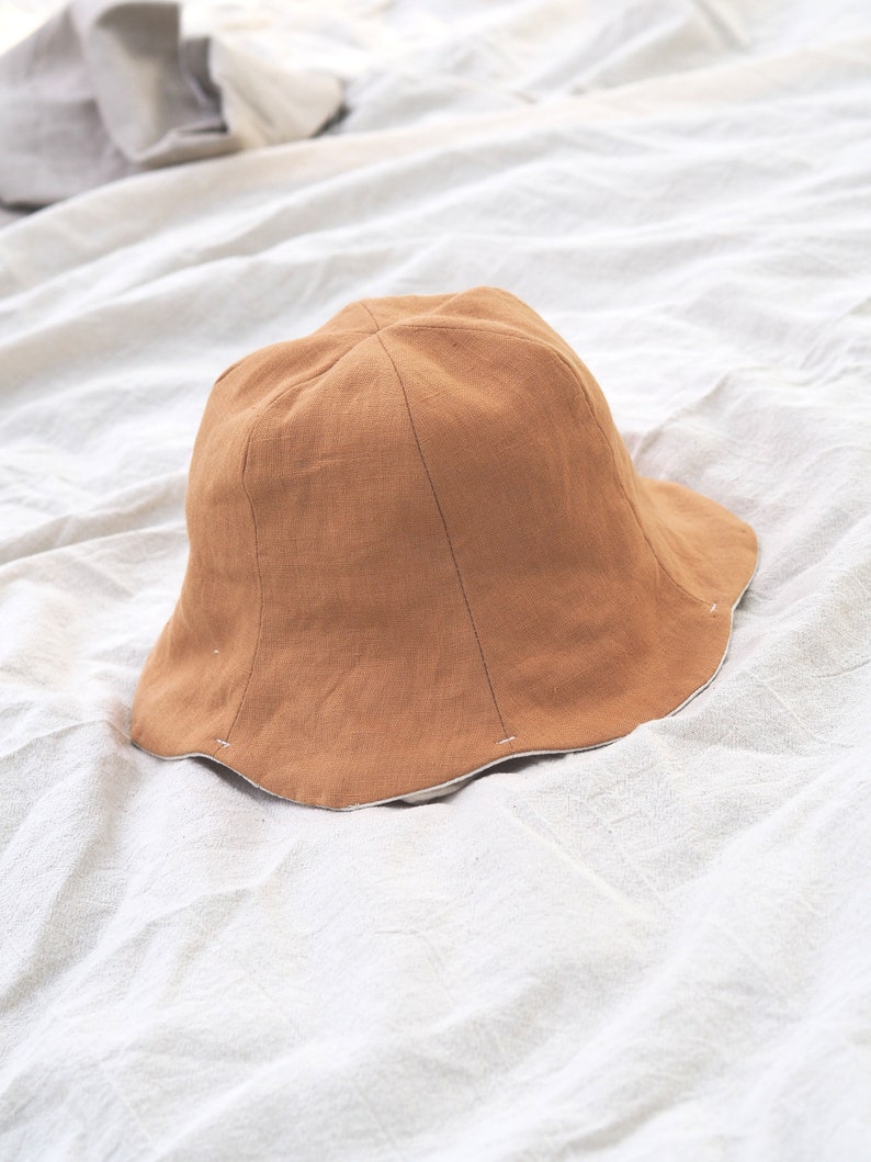 May include: A brown linen bucket hat with a white lining. The hat is sitting on a white fabric surface.