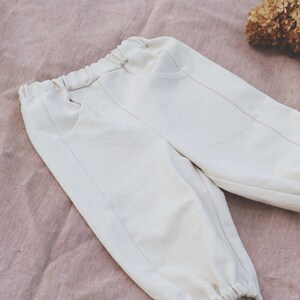 Op de afbeelding: Witte katoenen broek met een trekkoord in de taille en elastische boorden. De broek heeft twee zakken aan de voorkant.